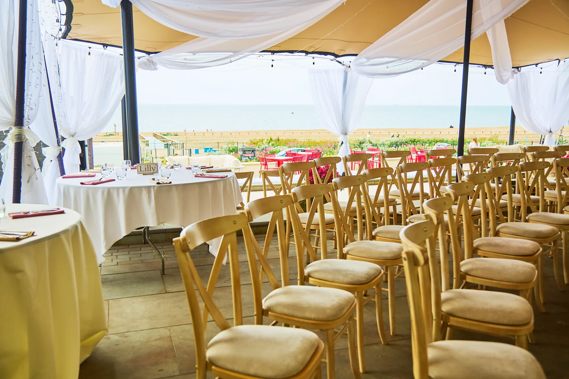 The terrace laid for a wedding: rows of cross-back chairs, round tables, white drapes and the sea beyond